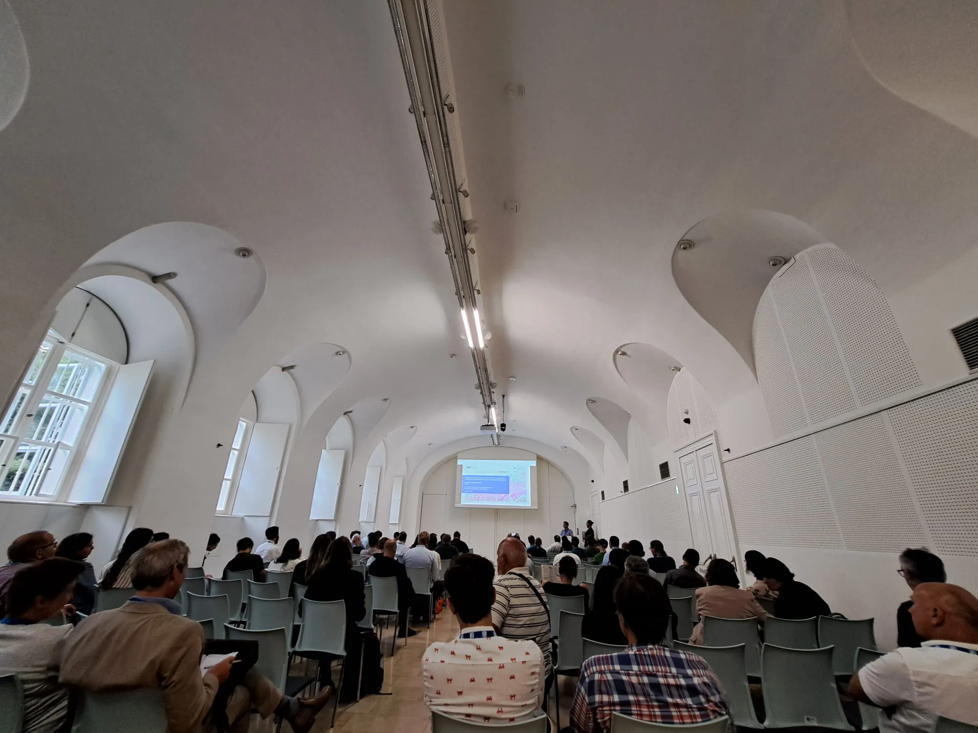  People seated in a bright conference room, watching a presentation.