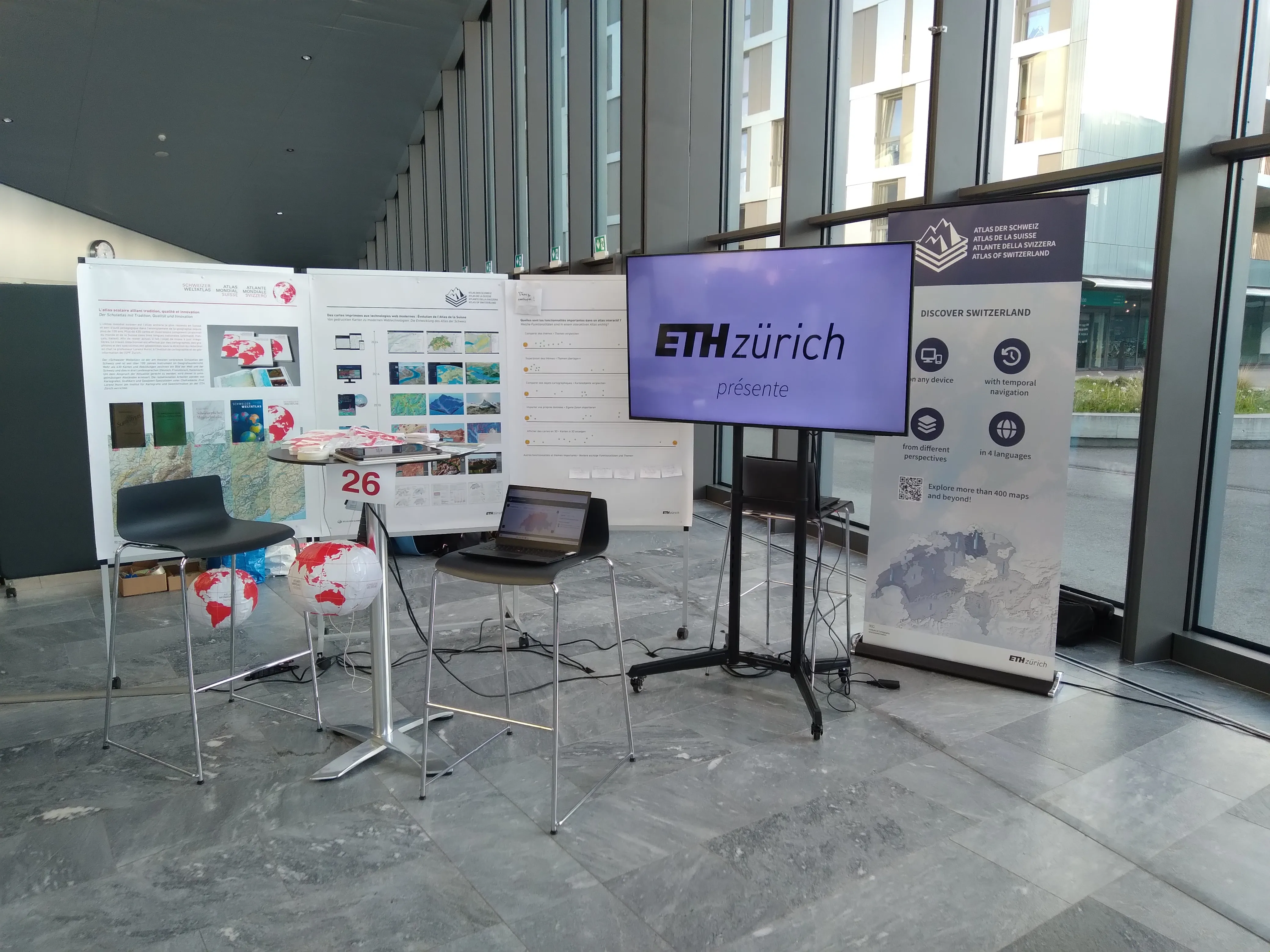 Ein Messestand über zwei Atlanten mit Bildschirm, drei Postern, einem Roll-up, zwei Stühlen und einem Computer.